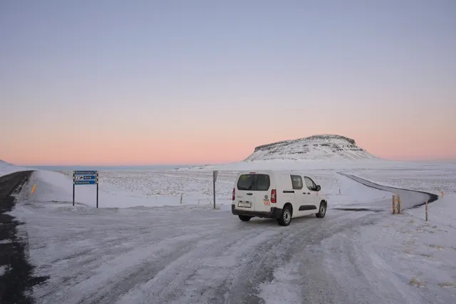 一台白色露營車停在冰島冬季的雪地公路旁，遠方是平頂雪山與淡粉色的天空