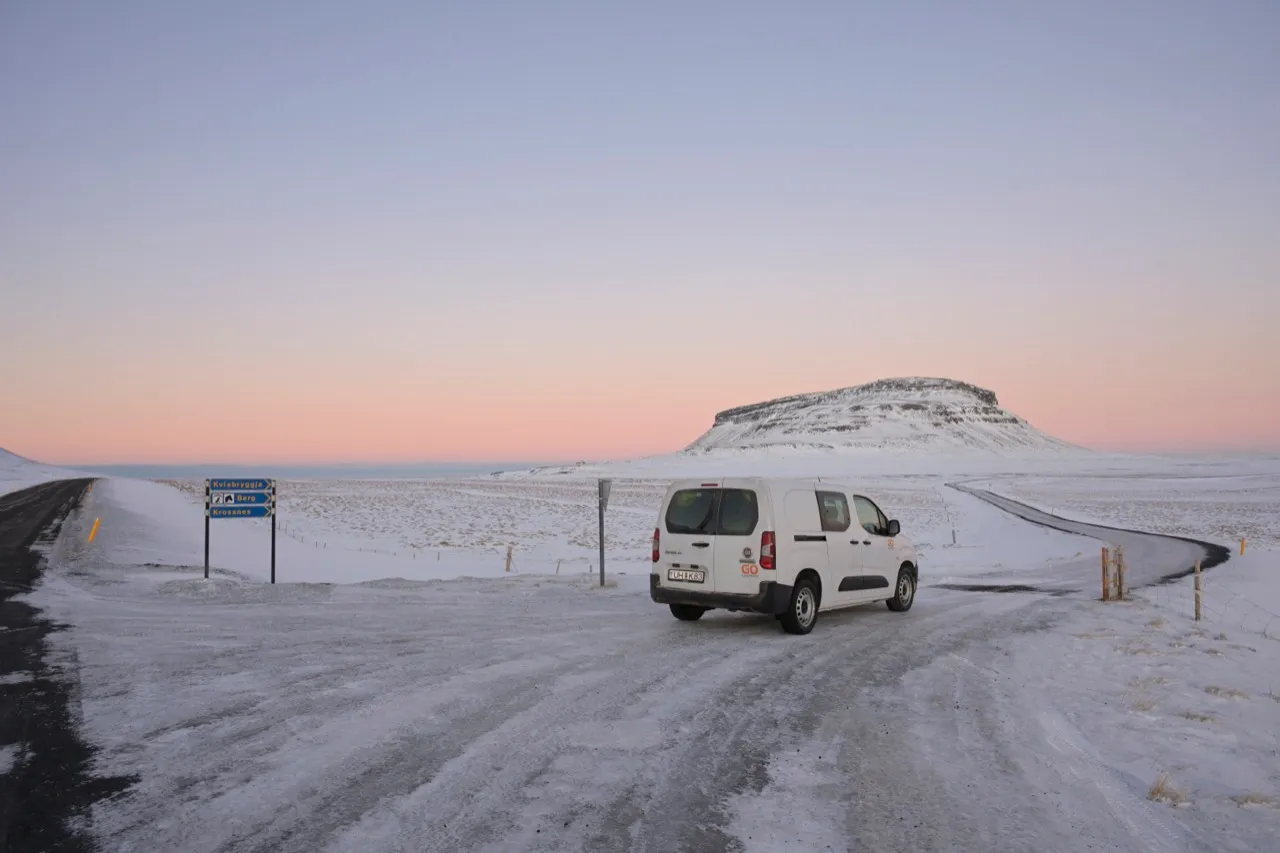 一台白色露營車停在冰島冬季的雪地公路旁，遠方是平頂雪山與淡粉色的天空