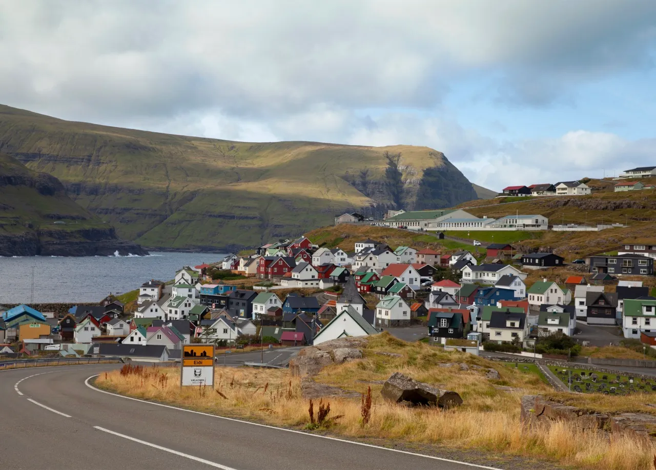 一條公路彎進法羅群島 Eiði 村，村裡彩色房舍沿著海灣分布，遠處是斷崖