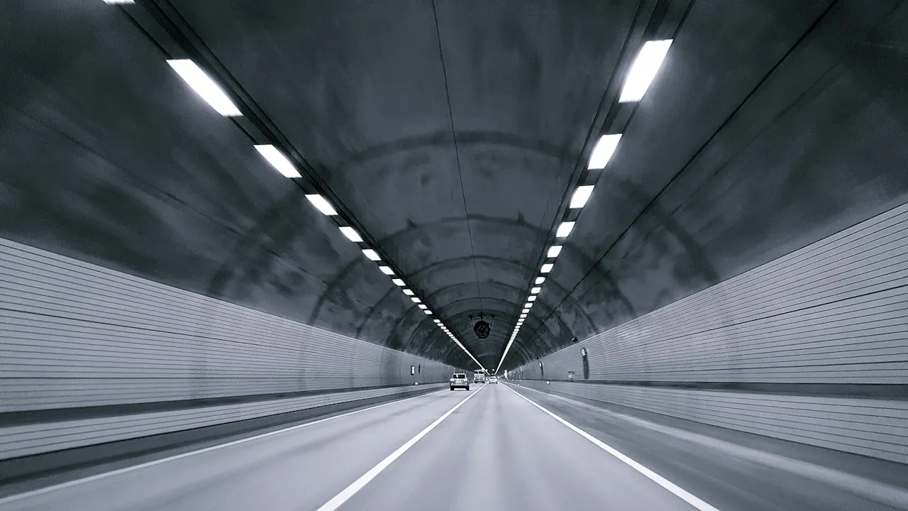 公路隧道內部，頂上連續燈帶延伸到遠處，路面畫著白色車道線