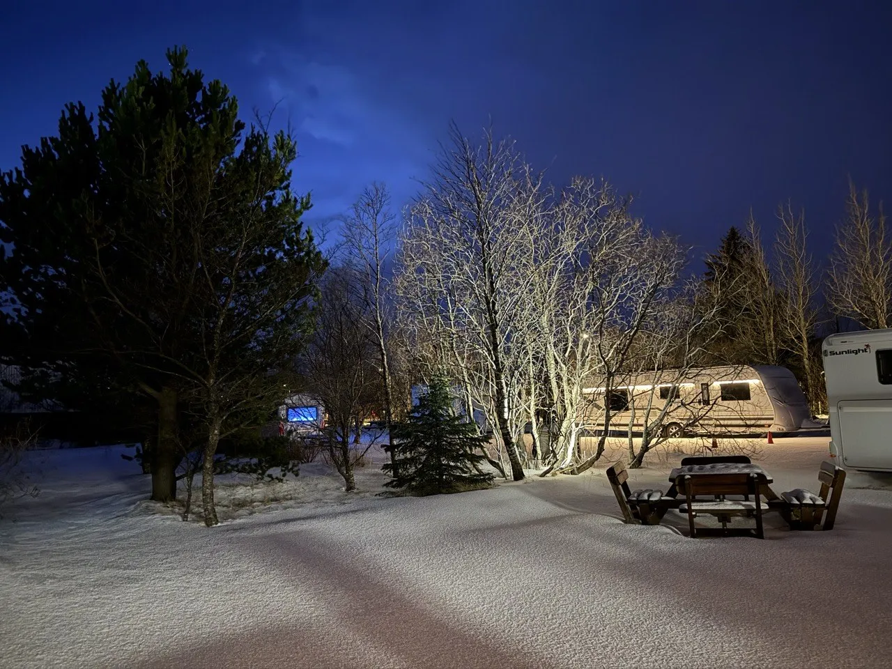夜間的冰島雪地營地,露營車停在樹林旁,遠處有微弱燈光